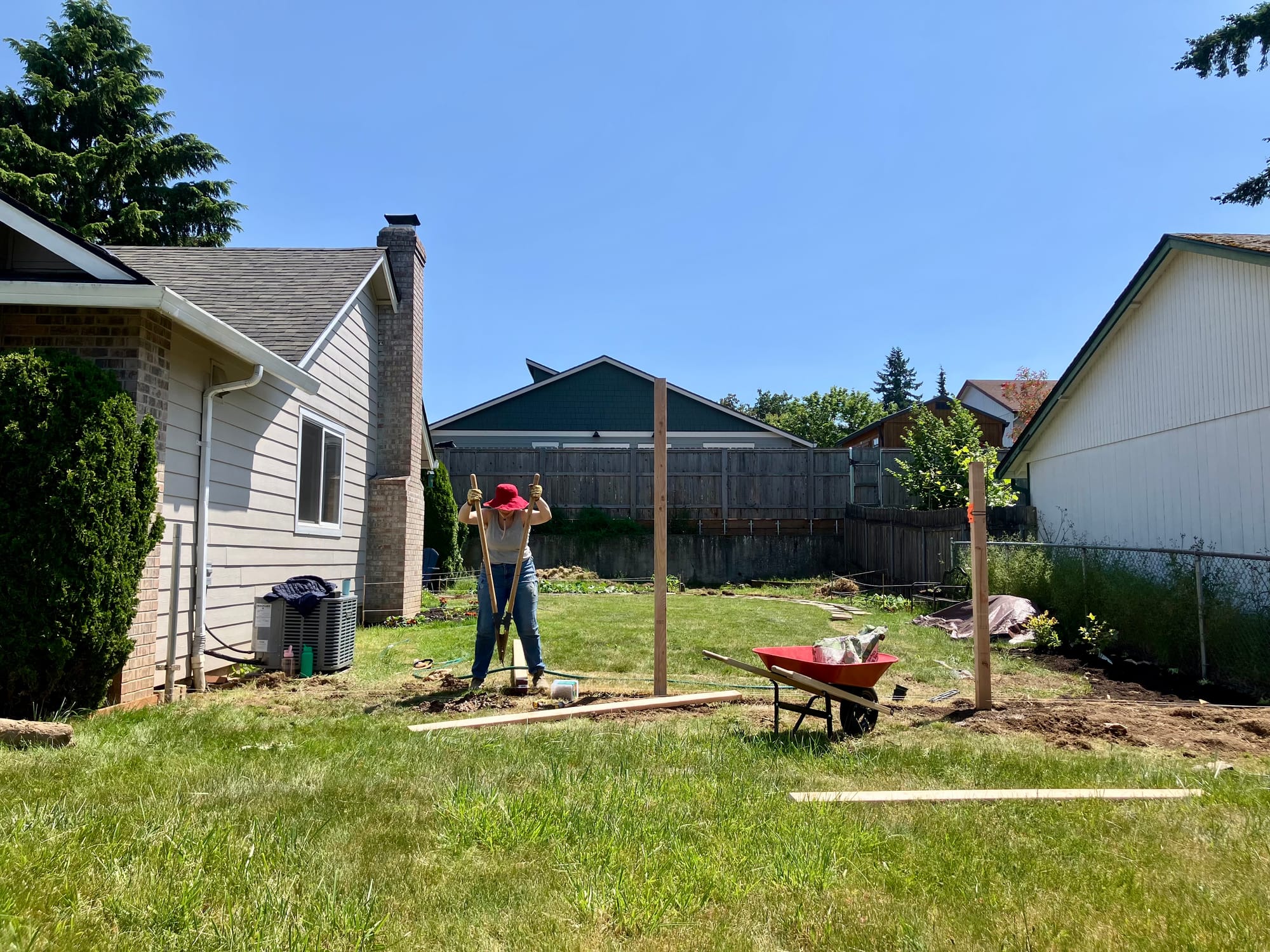 To the right of a beige house, a mom wearing a red hat uses a hole digger for a fence post
